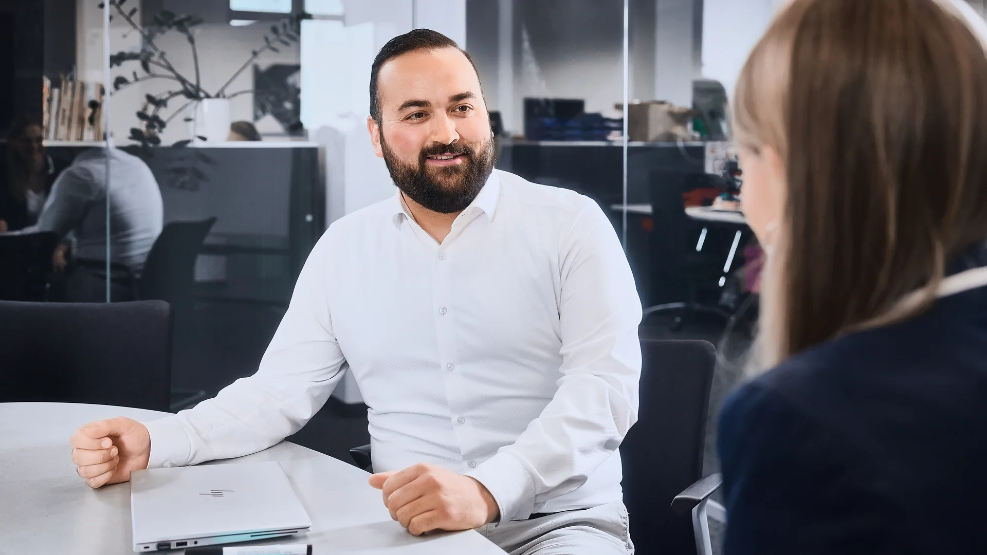 Ein Mann in weißem Hemd sitzt in einem Besprechungszimmer und unterhält sich mit der Frau gegenüber von ihm. 