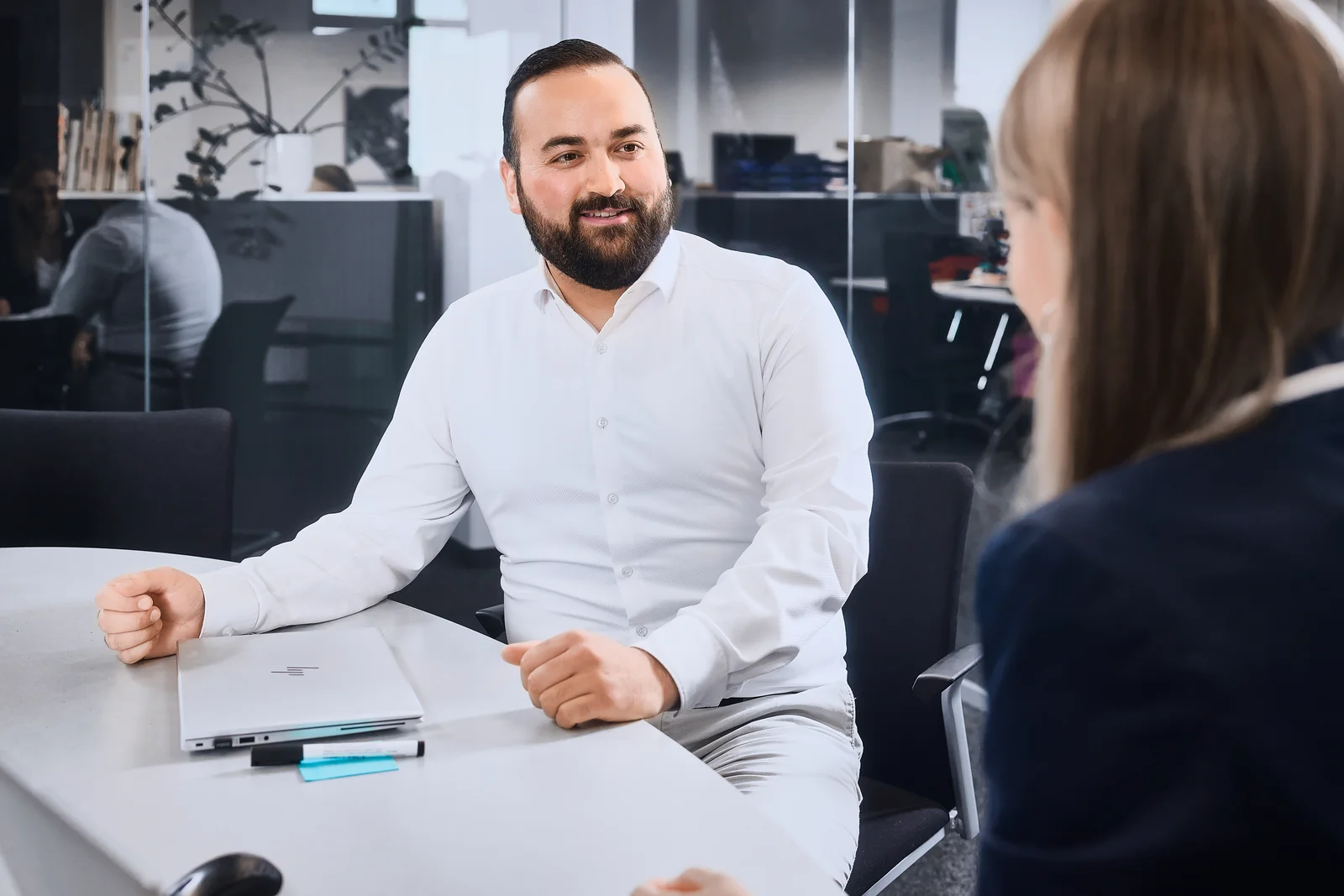 A man in a white formal shirt is sitting in a meeting room talking to the woman in front of him.