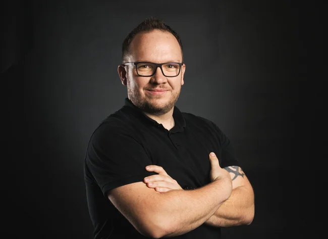 A man in a black T-shirt smiles at the camera and crosses his arms.