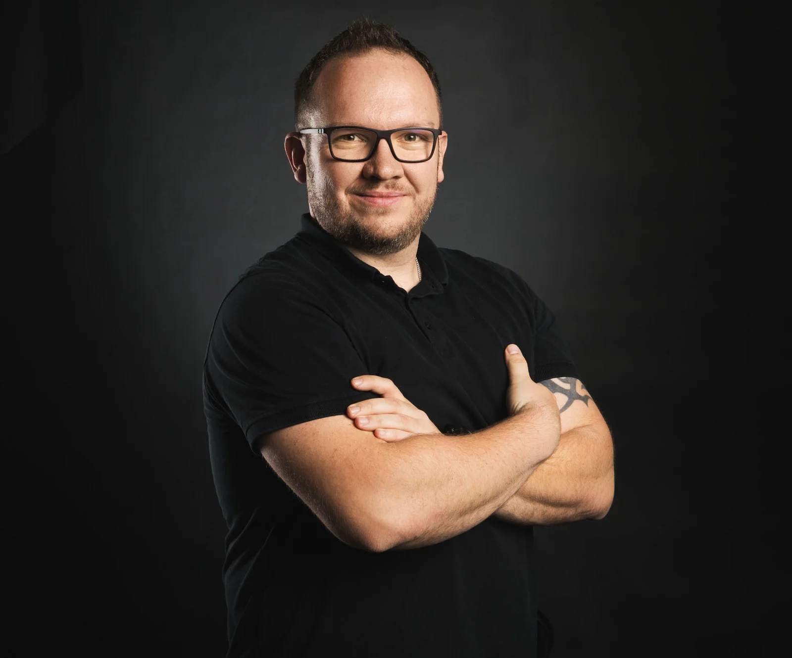 A man in a black T-shirt smiles at the camera and crosses his arms.