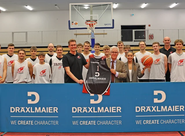 Die Baskets Vilsbiburg und der Head of Marketing der Firma DRÄXLMAIER stehen lächlend in der Basketballhalle.