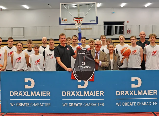 Die Baskets Vilsbiburg und der Head of Marketing der Firma DRÄXLMAIER stehen lächlend in der Basketballhalle.