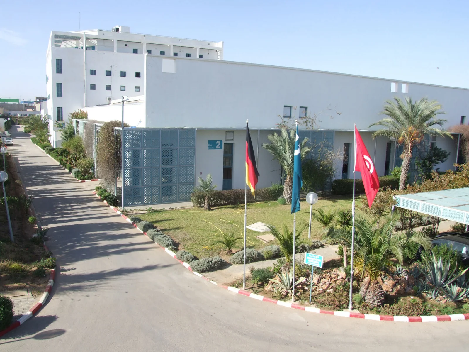 Weißes Gebäude mit mehreren Flaggen (Deutschland, DRÄXLMAIER, Tunesien) davor und einer Parkanlage mit Palmen in Sousse.
