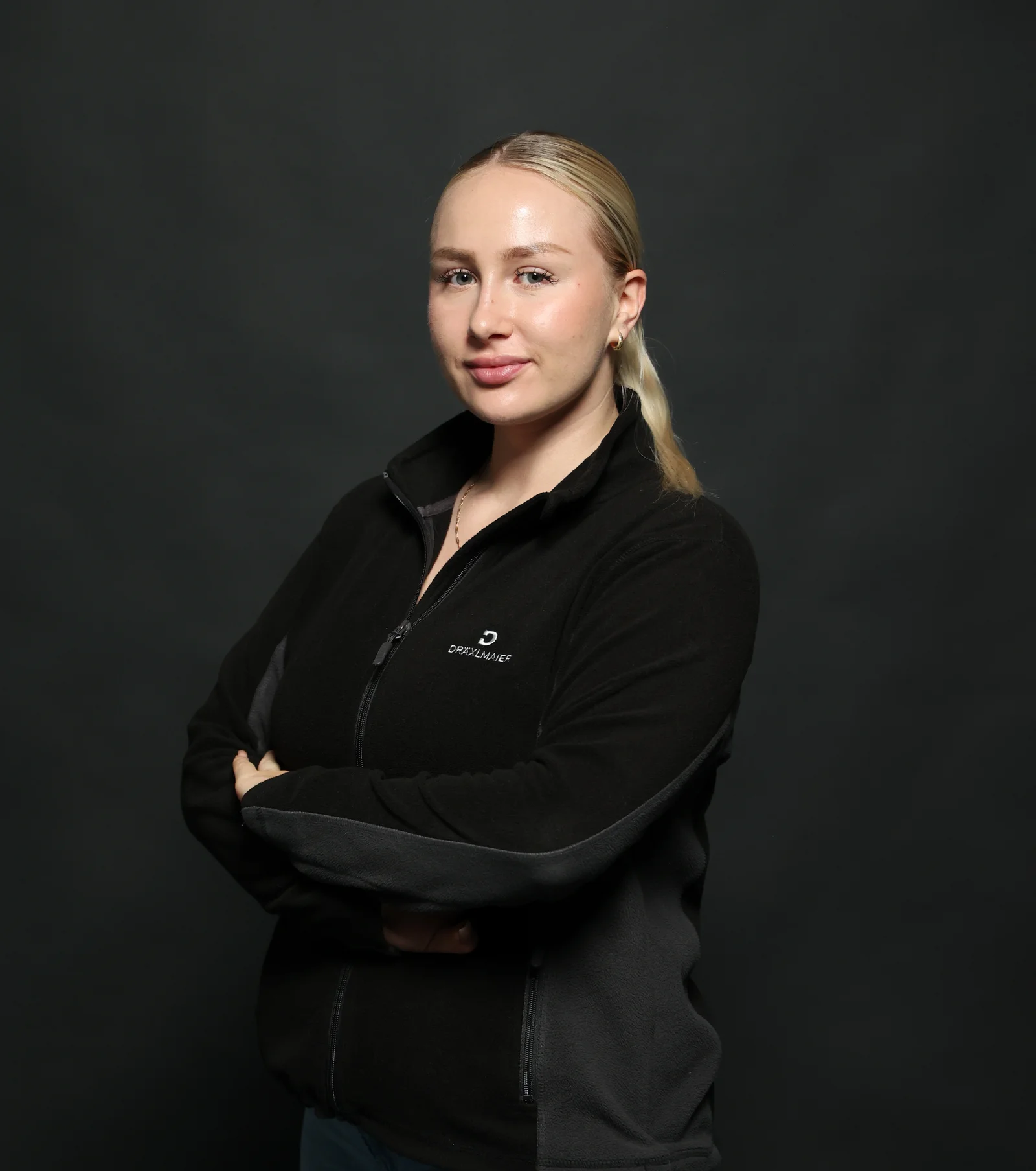 An apprentice wearing a black DRÄXLMAIER fleece jacket and looking into the camera with her arms crossed.