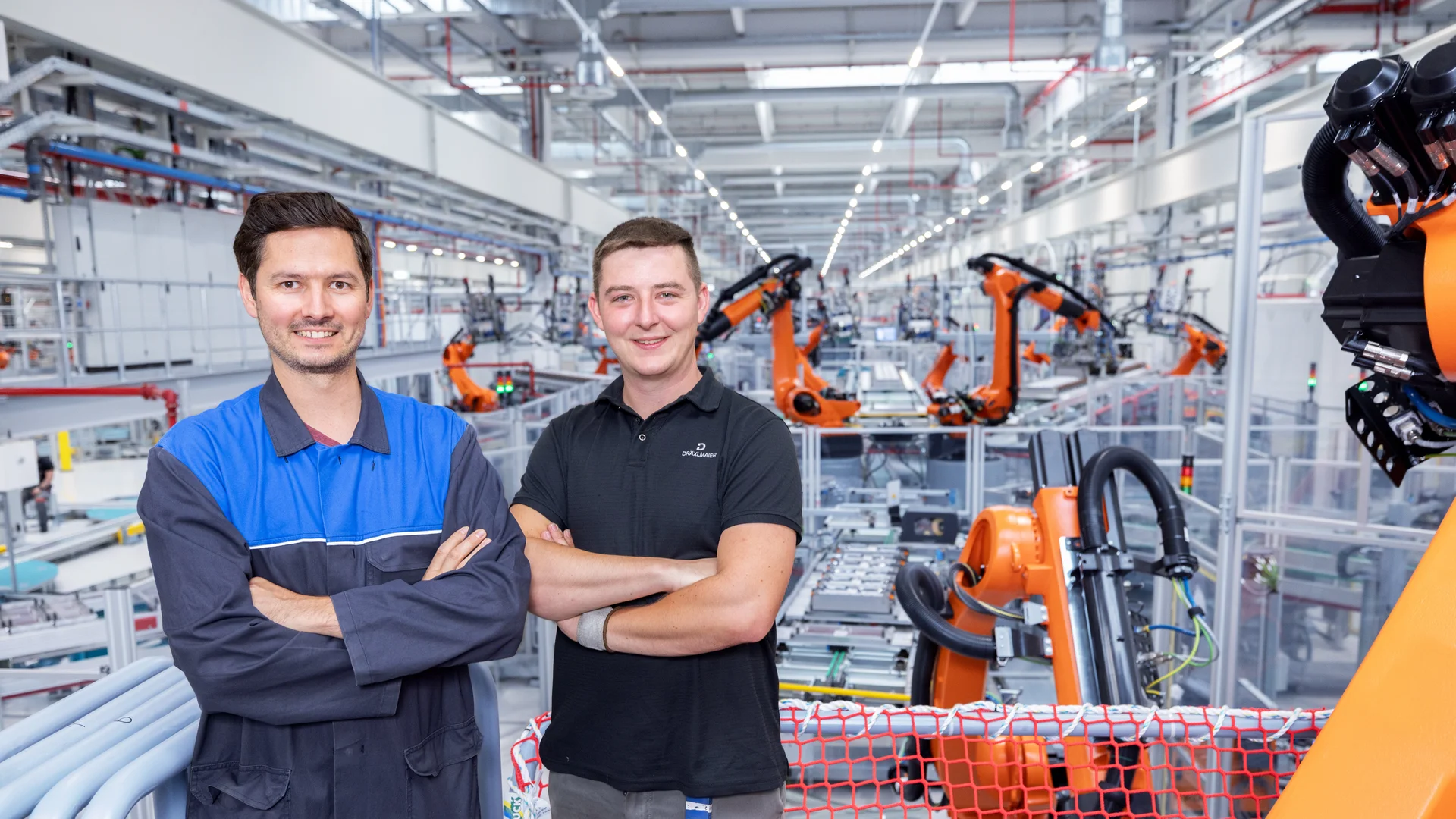 Zwei Mitarbeiter in einer DRÄXLMAIER Produktionshalle lächeln mit veschränkten Armen in die Kamera. 