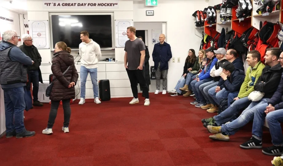Menschen in einem Hockey-Umkleideraum mit Ausrüstung, Bildschirm und dem Schild 'IT'S A GREAT DAY FOR HOCKEY'.