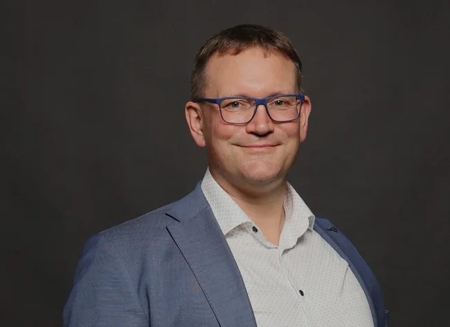 Ein Mann lächelt in die Kamera und hält seine Arme locker vor sich.