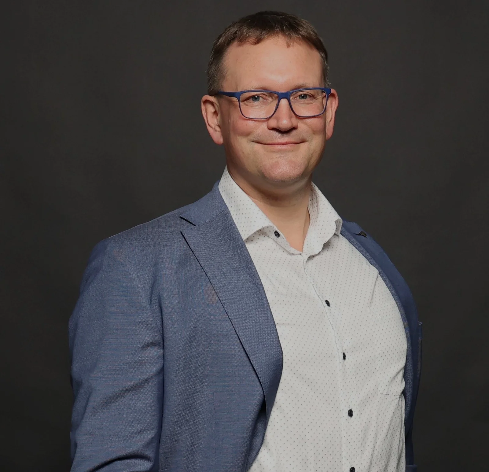 A man smiles at the camera and holds his arms loosely in front of him.