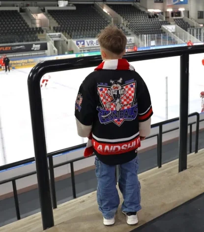 Person in einer ‚CANNIBALS LANDSHUT‘-Jacke schaut ein Eishockeyspiel in einer Halle mit Spielern in roten Trikots.