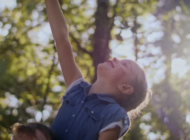 A girl on her father's shoulders. She stretches her arm upwards towards the treetops.