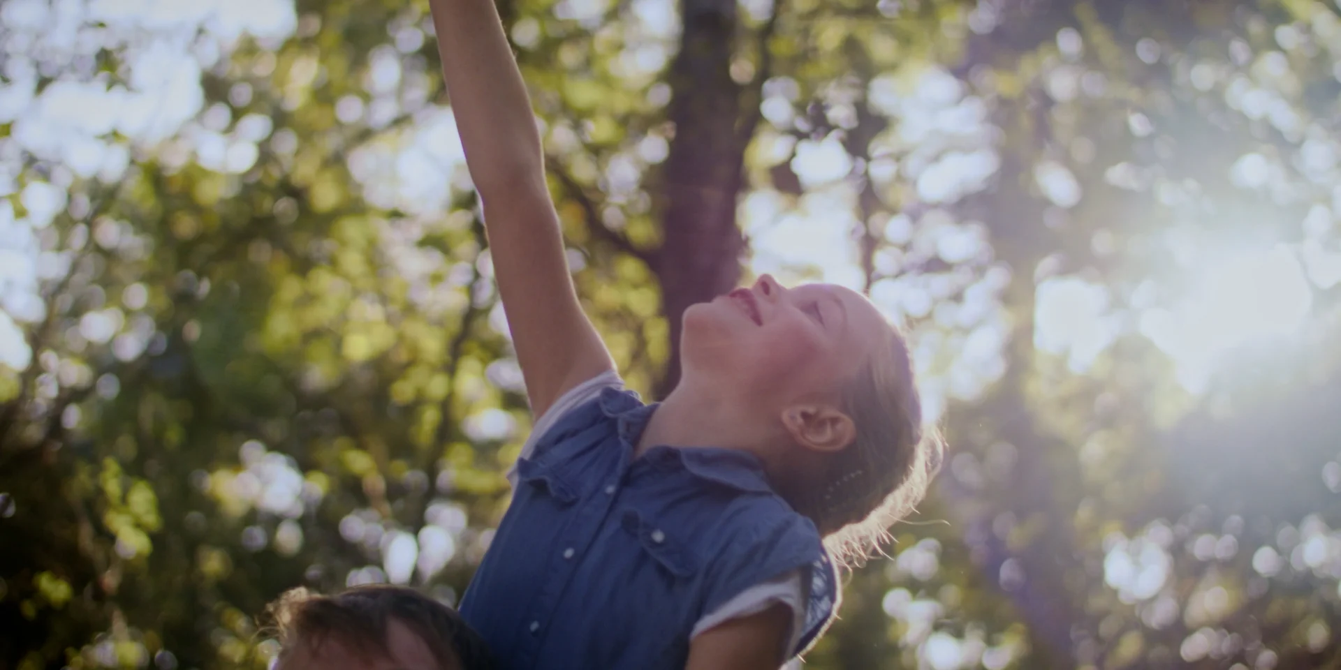 A girl on her father's shoulders. She stretches her arm upwards towards the treetops.