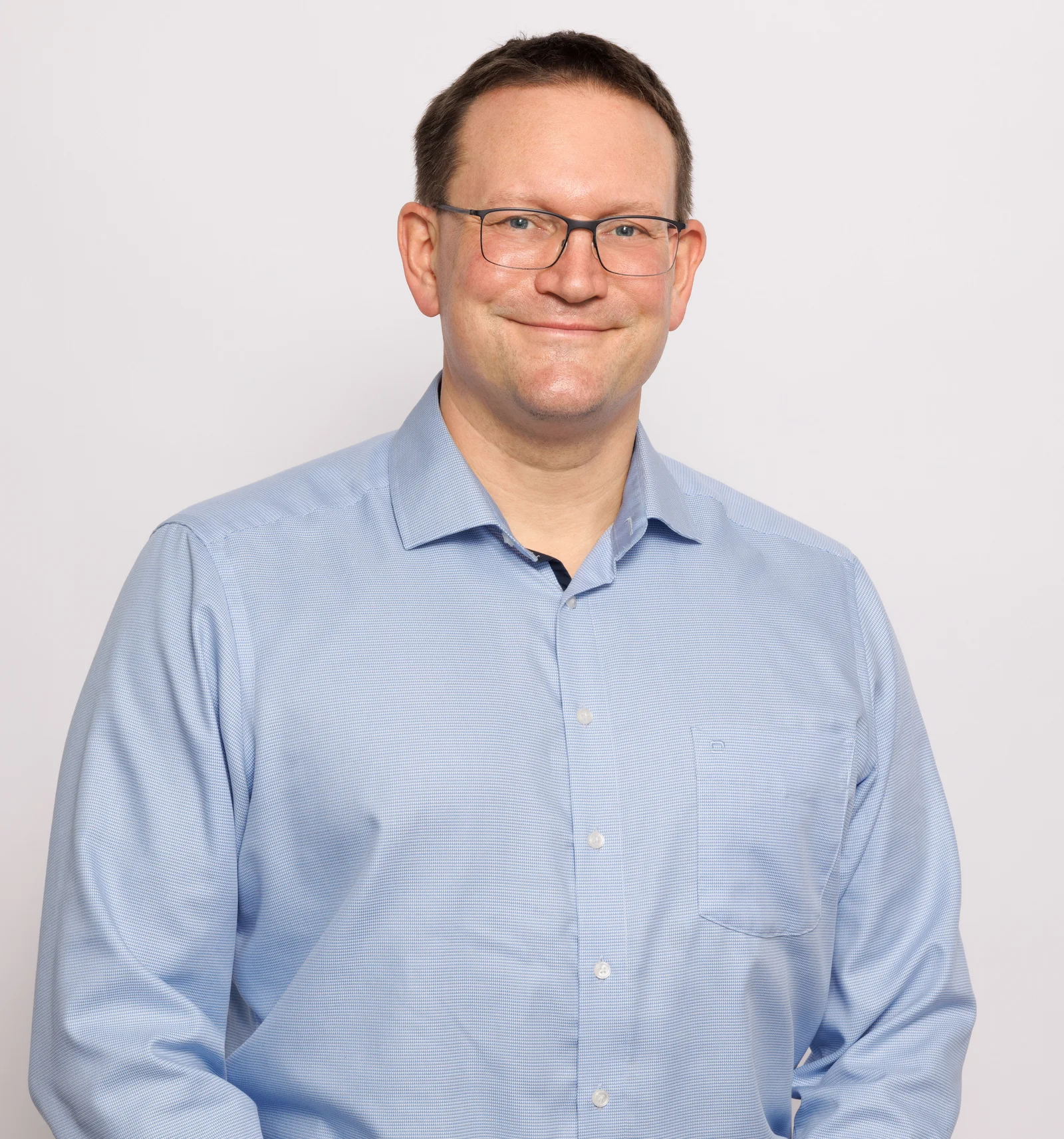 A man smiles at the camera and holds his arms loosely in front of him.