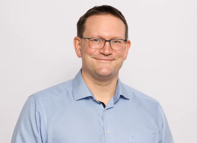 A man smiles at the camera and holds his arms loosely in front of him.