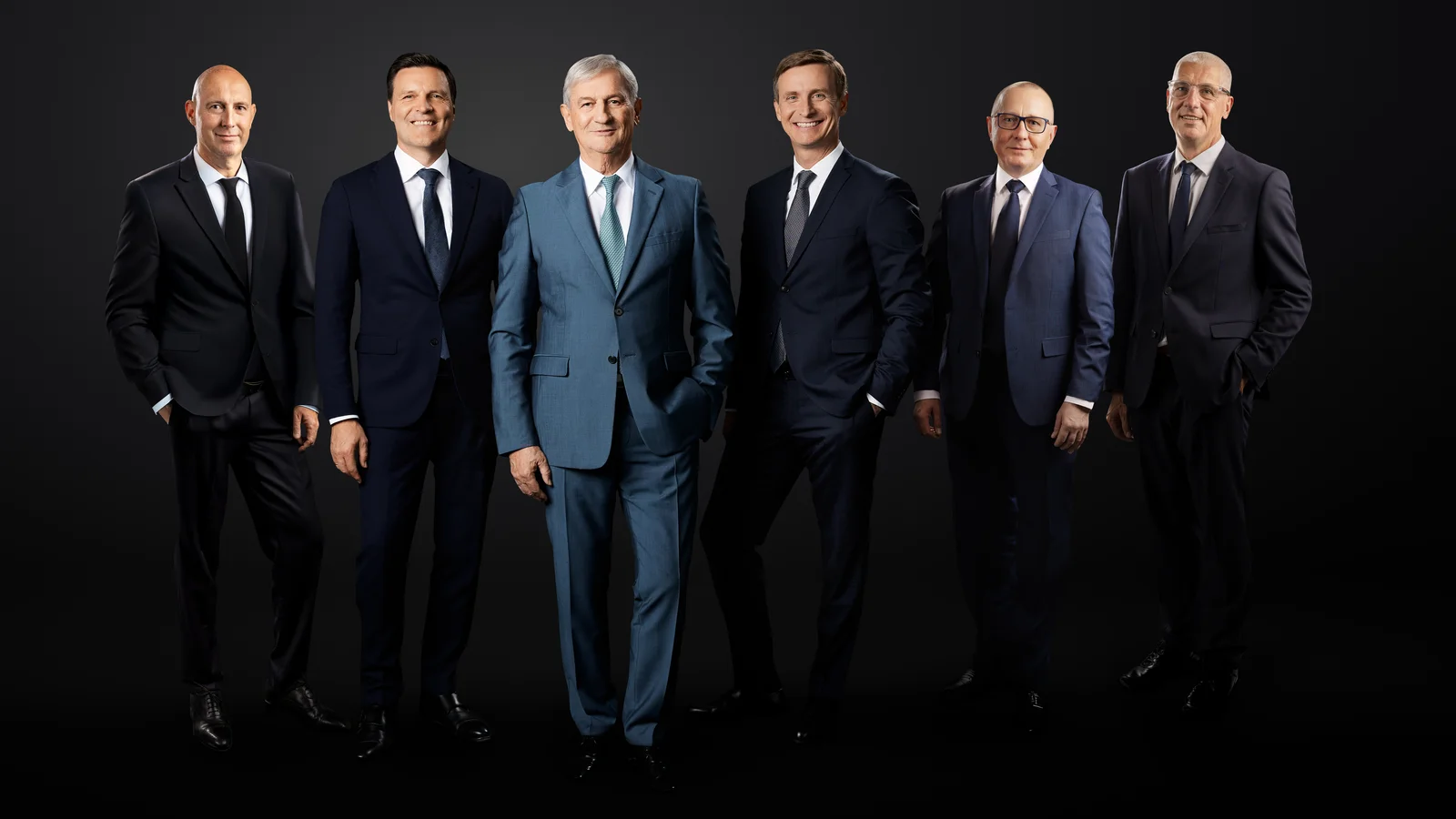 The main management in front of a black background. Six men smile into the camera. 