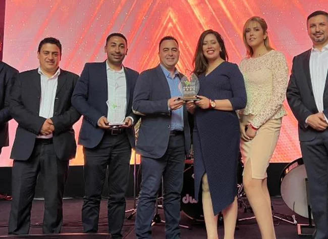 Seven people stand in a row and smile at the camera. Two people in the middle are holding the award. 