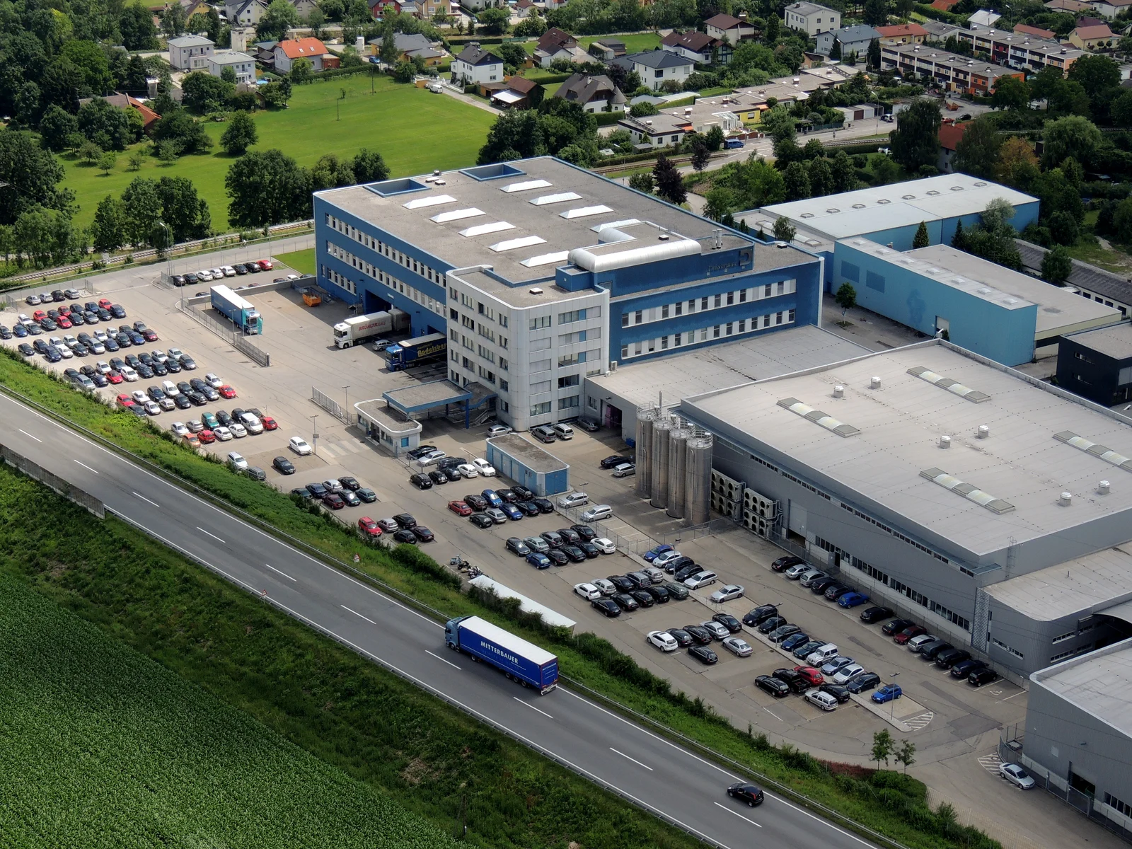 Luftaufnahme des großen Industriekomplexes in Braunau mit mehreren Gebäuden und vielen parkenden Autos.