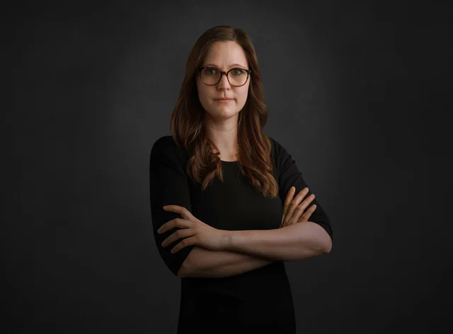 A woman with glasses stands with her arms crossed in front of a black background.