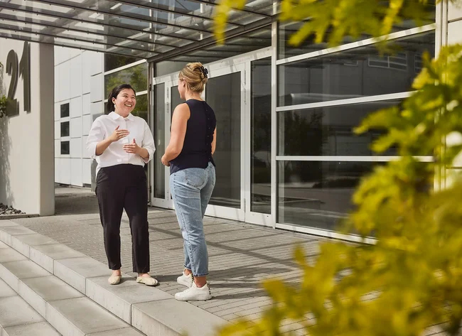 Zwei Frauen stehen vor einem DRÄXLMAIER Geäude und unterhalten sich. 
