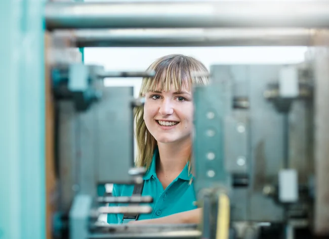 Eine Auszubildende zur Verfahrensmechanikerin lächelt in die Kamera. Im Vordergrund erkennt man die gefertigten Werkstücke.