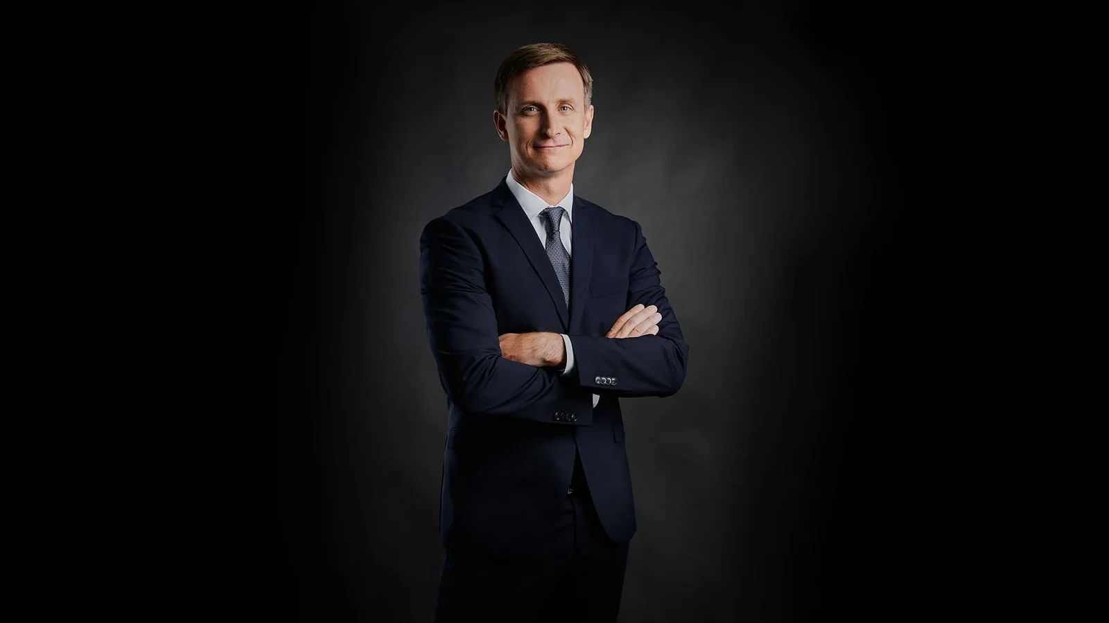 A man stands smiling with his arms crossed in front of a black background.