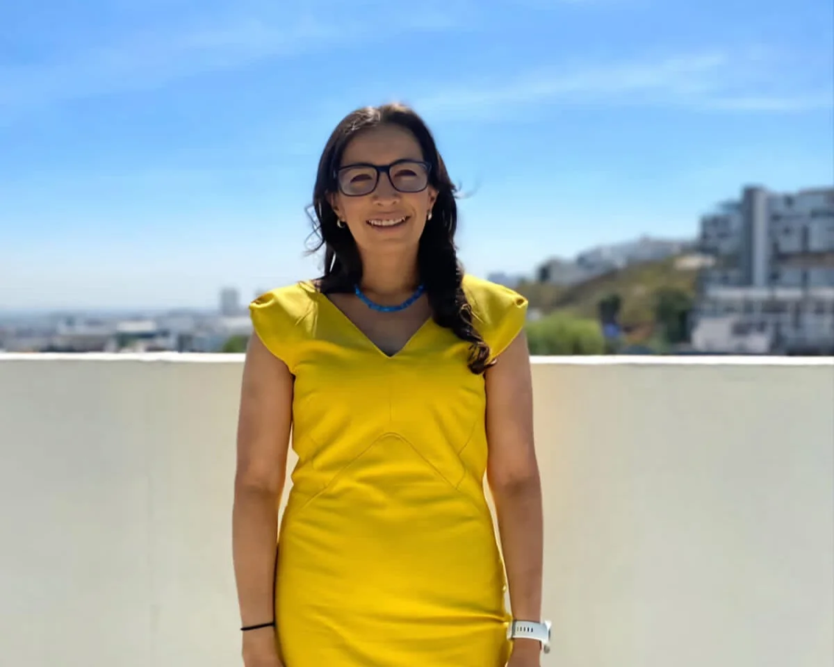 A woman in a yellow dress smiles at the camera. Blue sky and houses in the background.
