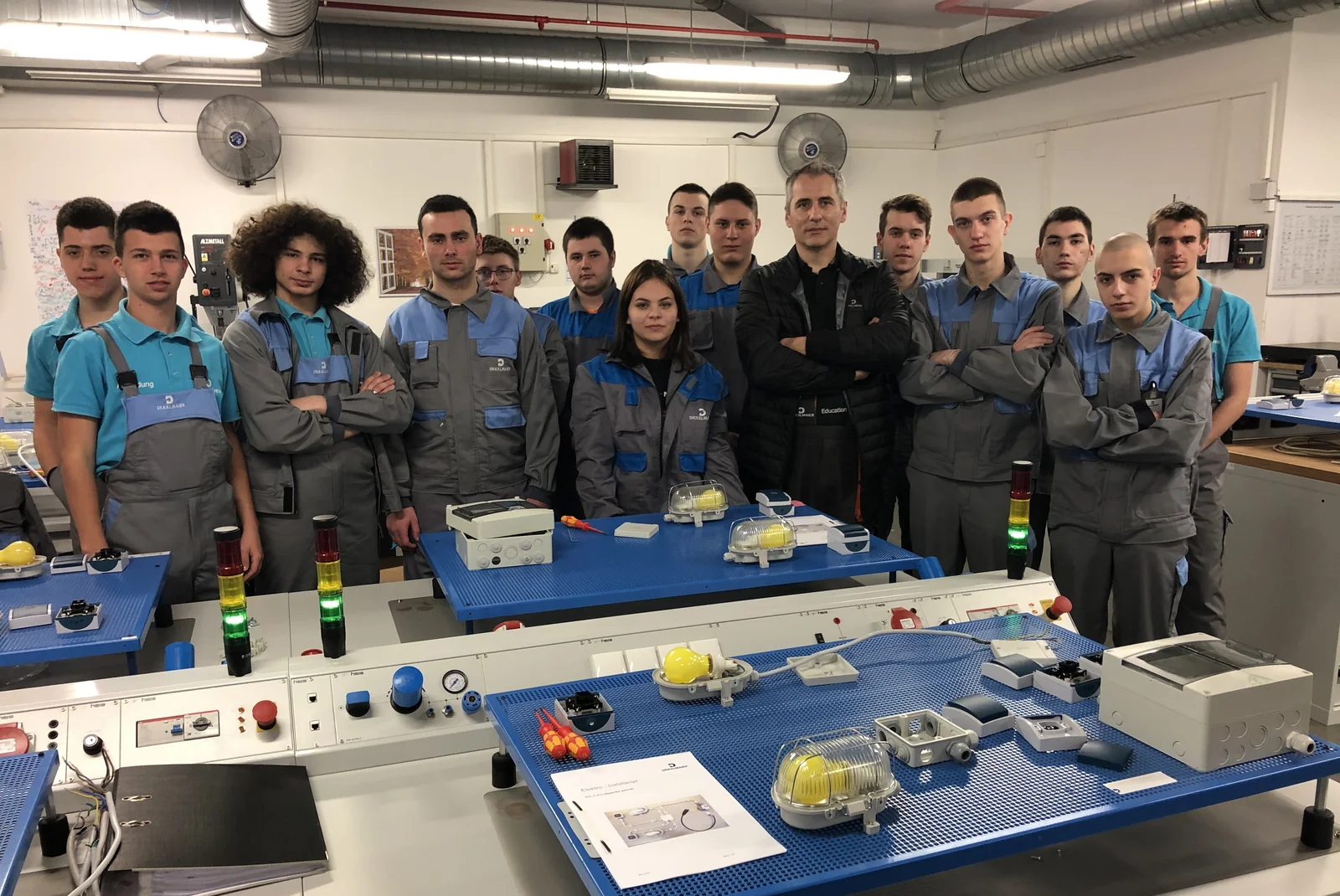 Ein Ausbilder steht mit vierzehn Auszubildenden in der Lehrlingswerkstatt für Elektronik. Alle schauen in die Kamera.