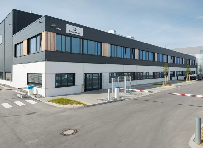 The company building in Sachsenheim with a grey façade and wooden cladding, a barrier and a zebra crossing in front of it.