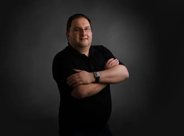 A man with glasses stands with his arms crossed in front of a black background.