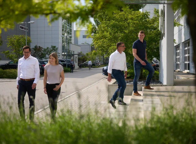 Four people talking on the DRÄXLMAIER premises. Green trees in the background.