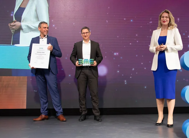 Zwei Männer und eine Frau lächeln in die Kamera. Ein Mann hält die BME Auszeichnung. Der andere Mann hält den BME Award. 