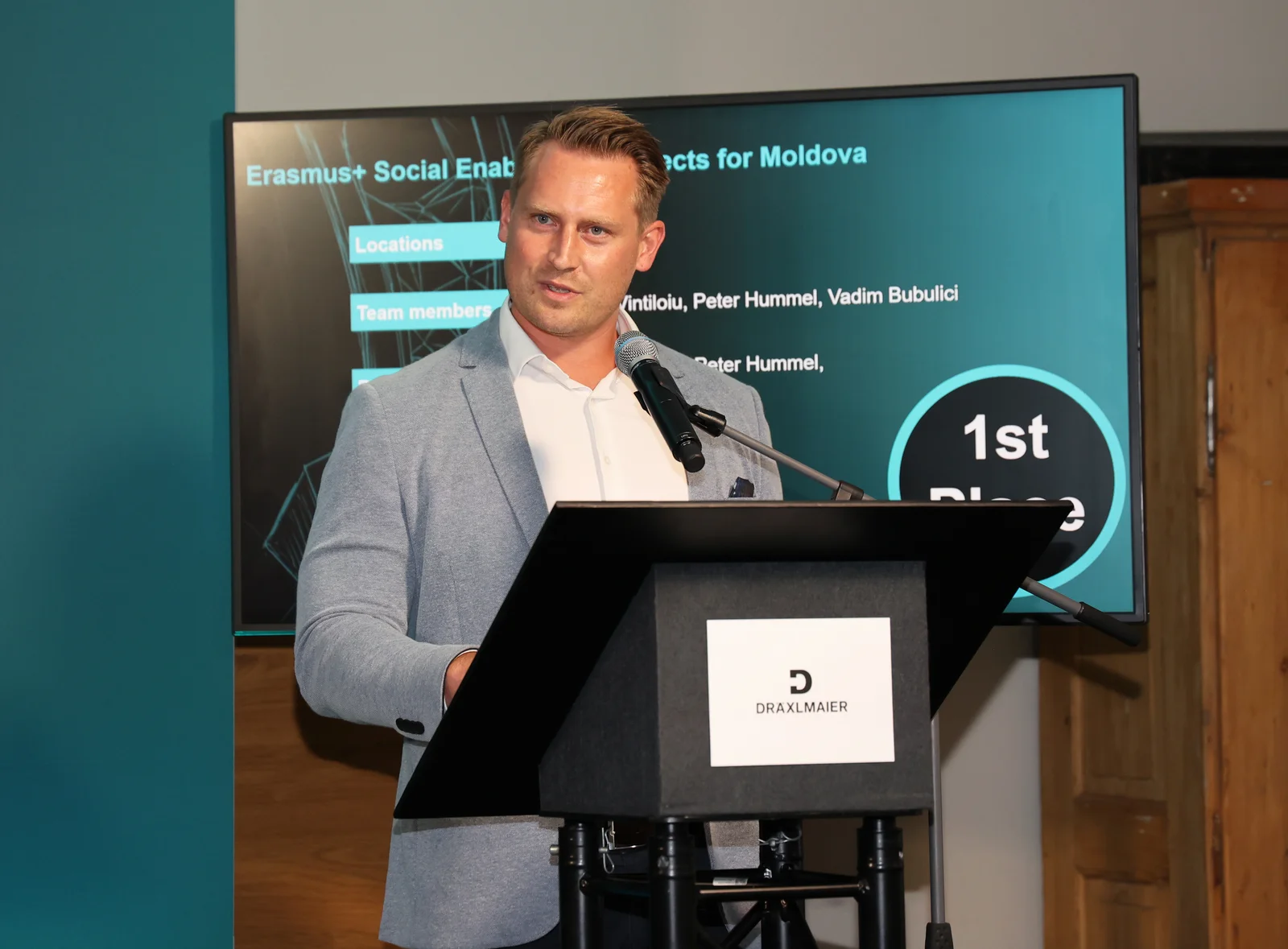 A man is giving a speech at a DRÄXLMAIER lectern. There is a large screen in the background.