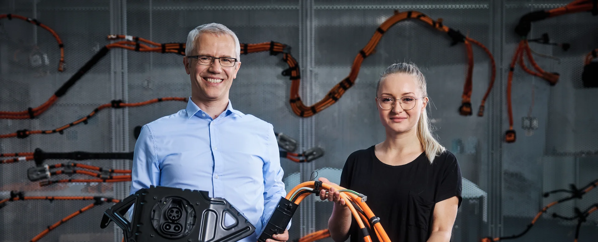 A man and a woman smile at the camera. They are holding part of a cable harness in their hands.