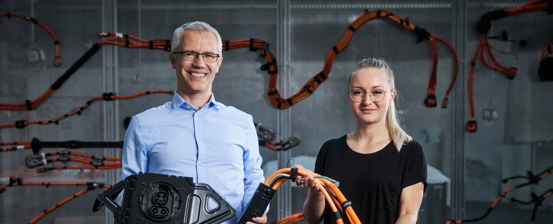 Ein Mann und eine Frau lächeln in die Kamera. Sie halten einen Teil eines Kabelbaums in den Händen.