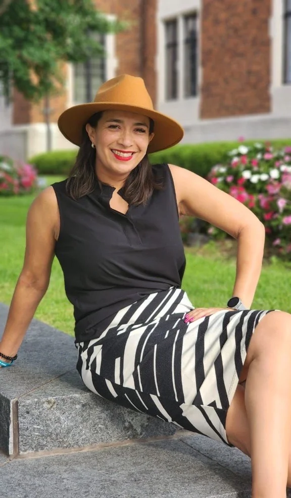 Woman with hat and dress sits on a stone with her legs crossed.