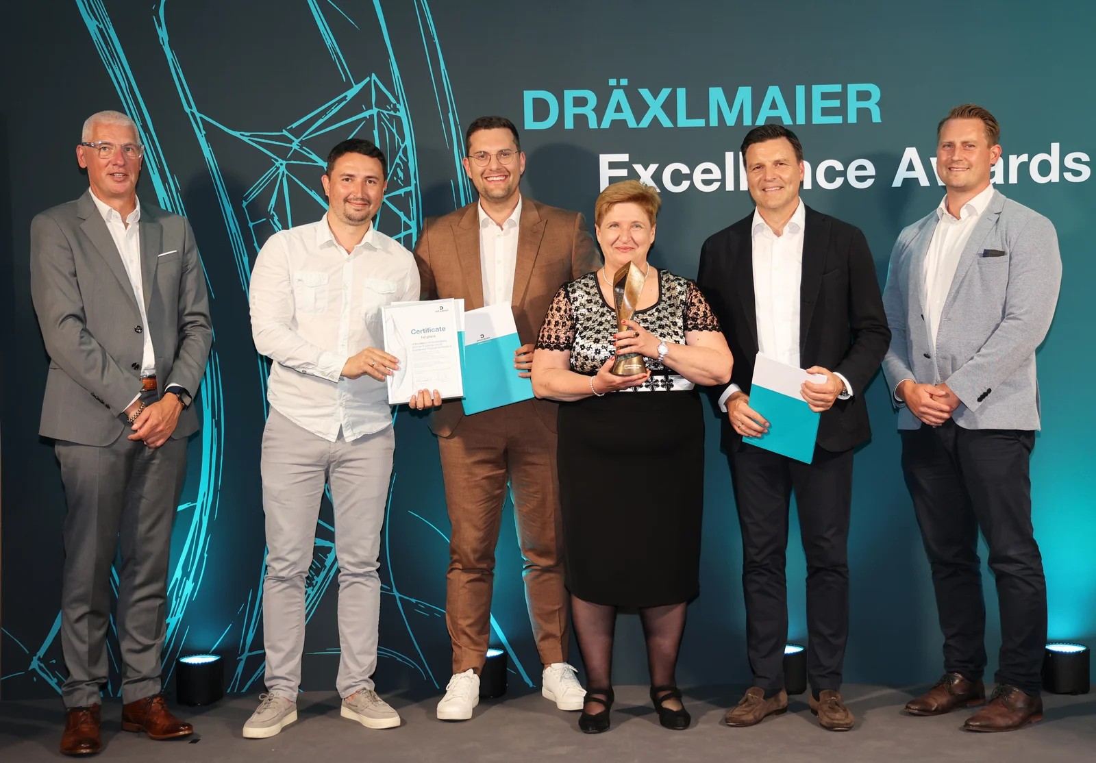 Six people stand in front of a screen labelled ‘DRÄXLMAIER Excellence Awards’. They are holding certificates and a trophy.