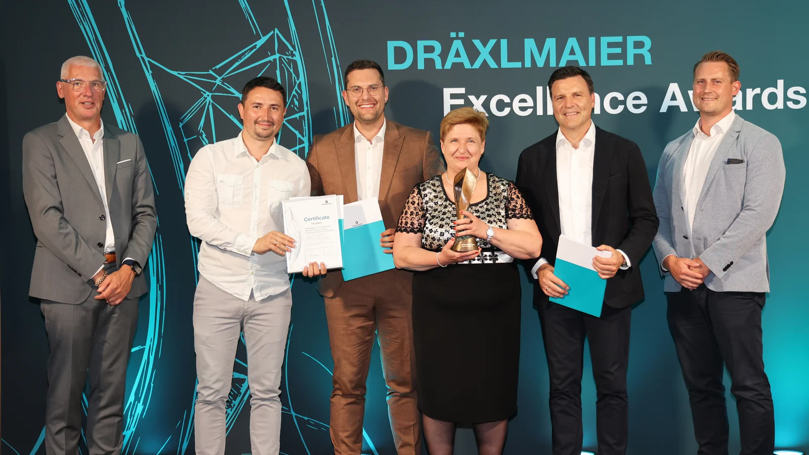 Six people stand in front of a screen labelled ‘DRÄXLMAIER Excellence Awards’. They are holding certificates and a trophy.