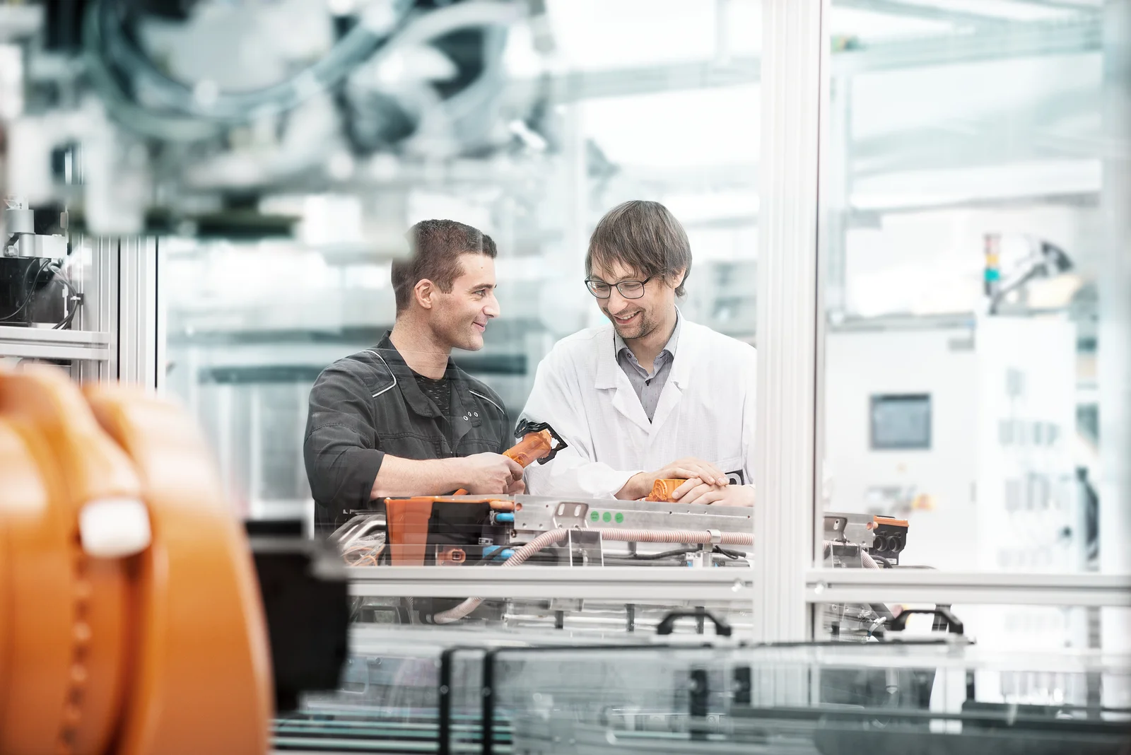 Zwei Personen in Arbeitskleidung stehen in einer industriellen Umgebung und arbeiten an Maschinen.