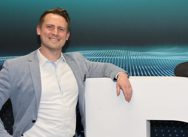 Ein großes weißes DRÄXLMAIER "D" steht im Vordergrund. Zwei Männer sützten sich mit den Armen ab und lächeln in die Kamera. 