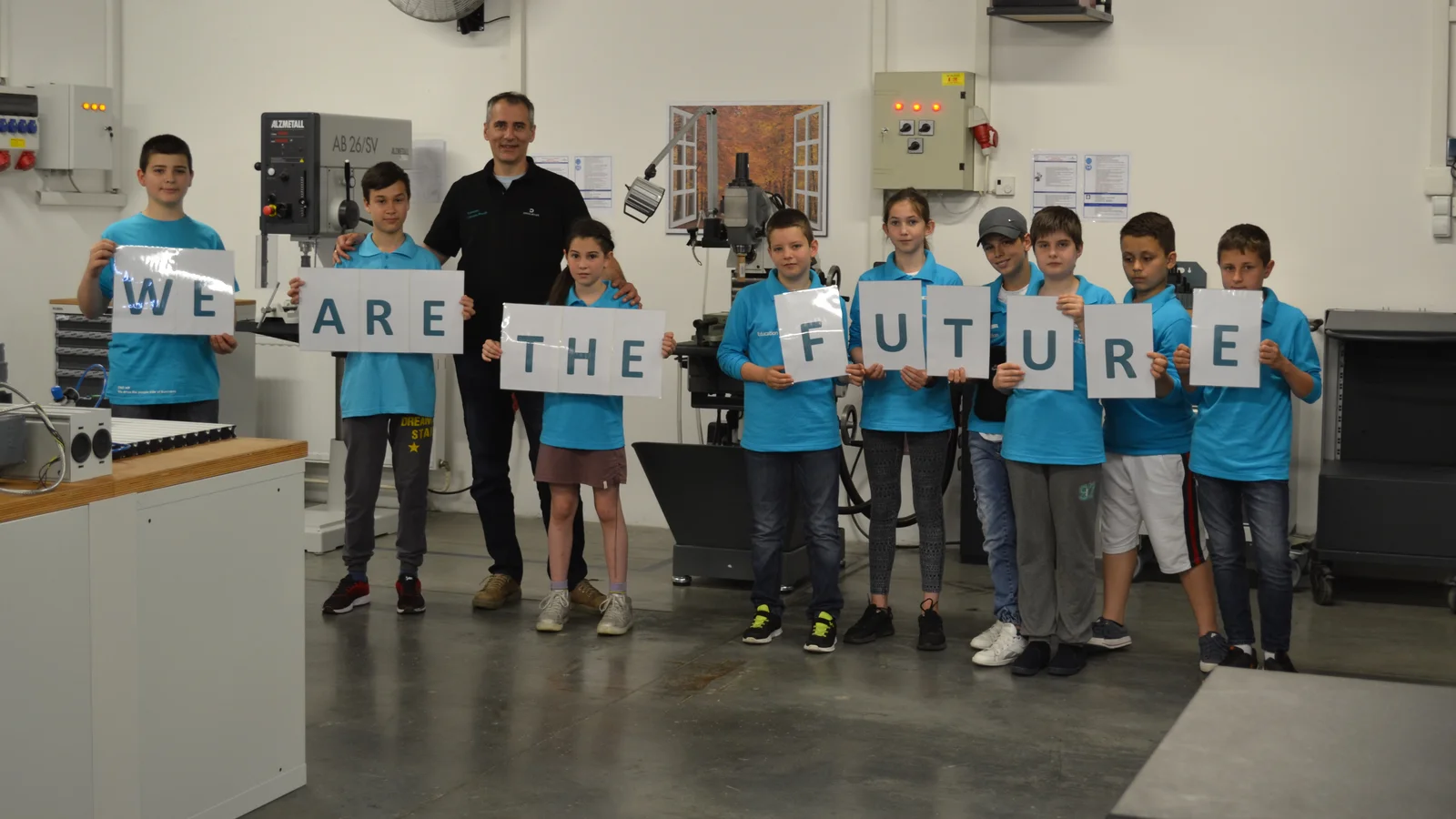 Ein Ausbilder mit 9 Kindern in einer Werkstatt. Die Kinder halten Buchstaben fest, die den Satz "We are the future" ergeben.