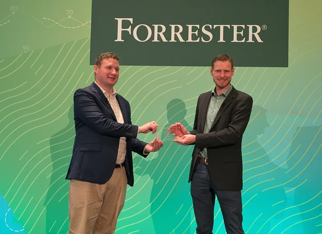 Two people stand in front of a green wall. Together they are holding the London Forrester Award. 