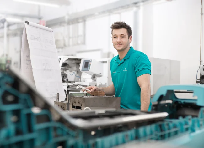 Ein Auszubildender in der Werkzeugmechanik bei DRÄXLMAIER in Vilsbiburg lächelt und arbeitet an einem Werkstück.