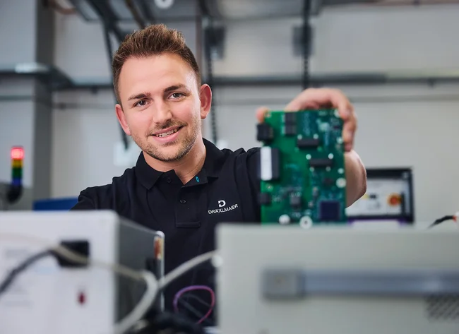 DRÄXLMAIER employee smiles at the camera and holds a green circuit board. 