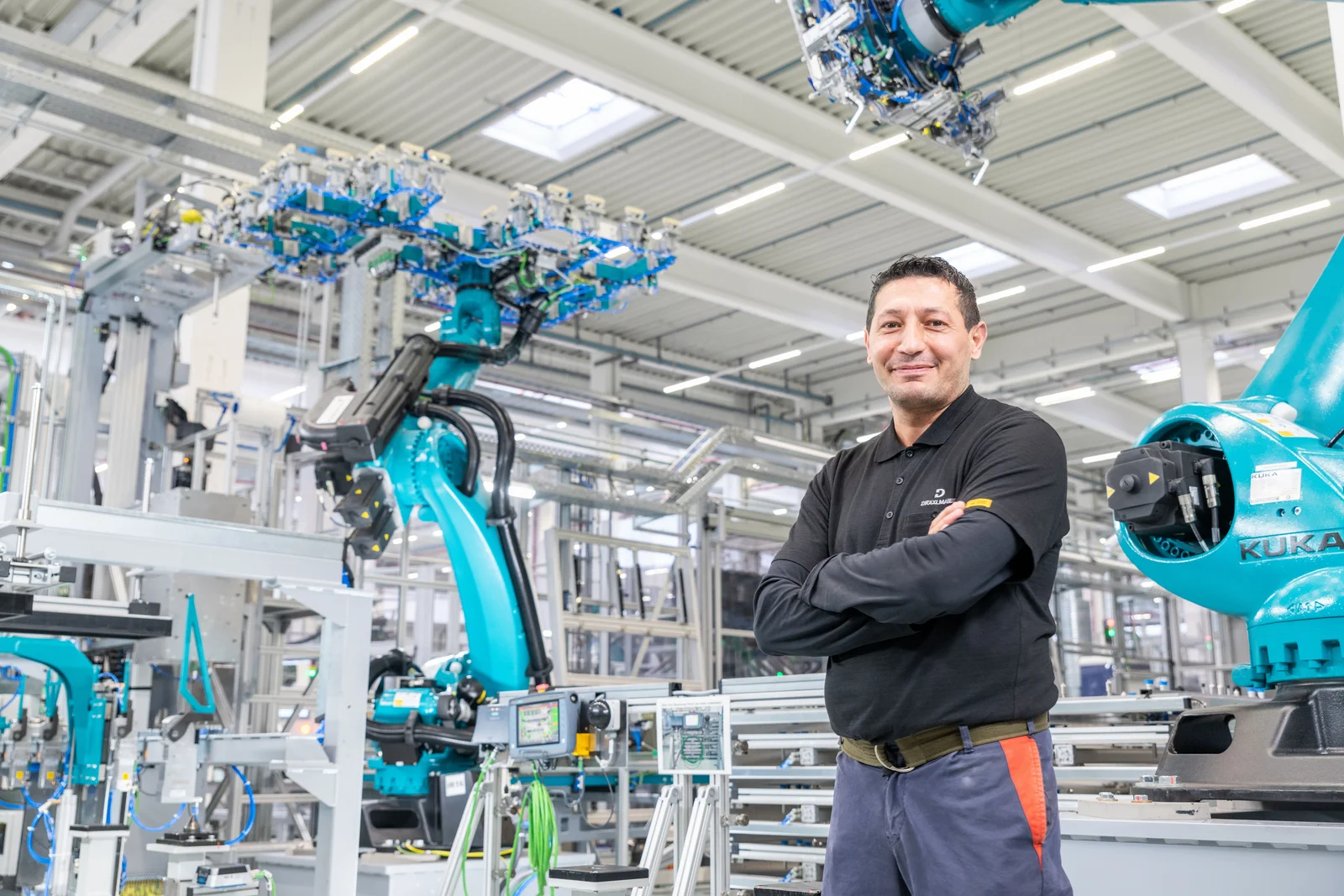 Ein Mann steht mit verschränkten Armen in einer Fabrikhalle, umgeben von blauen Industrierobotern und Maschinen.