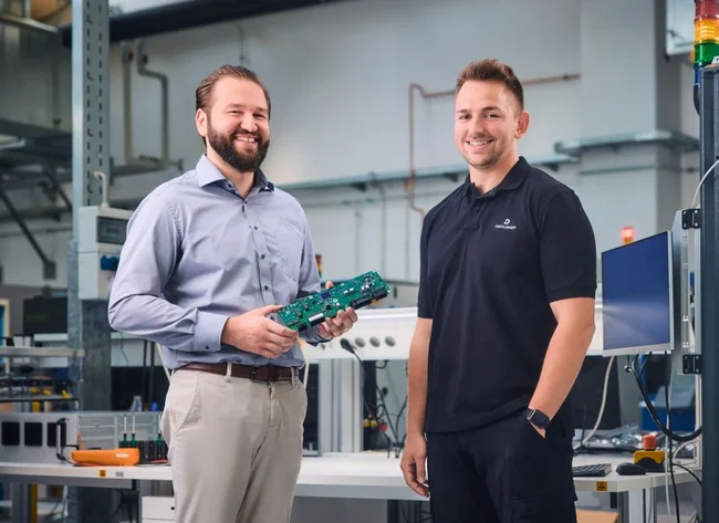 Two men, one in a formal shirt, one in a DRÄXLMAIER T-shirt, smile at the camera.