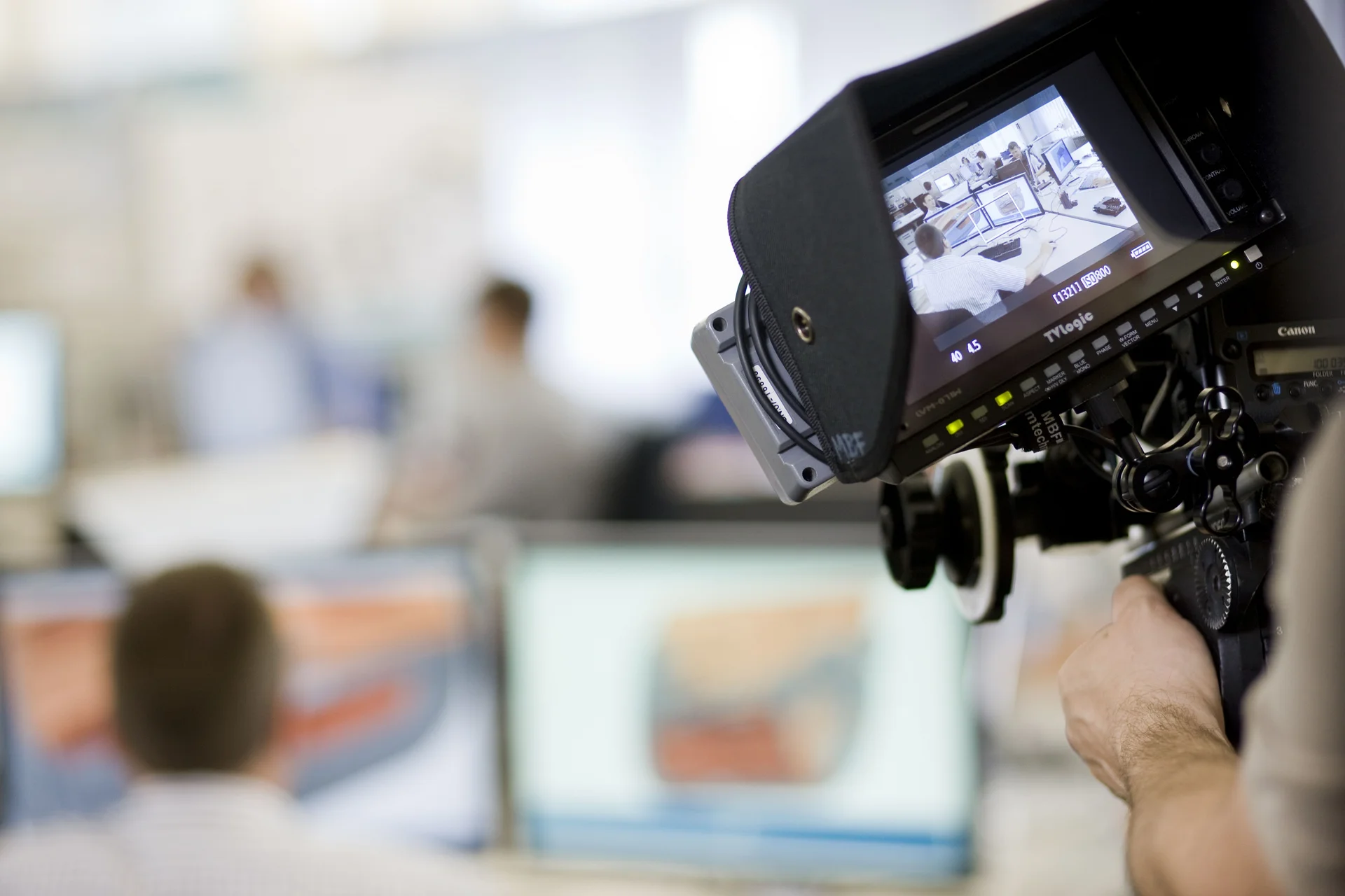 A camera films people in front of a computer screen. The background is blurred.