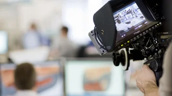 A camera films people in front of a computer screen. The background is blurred.
