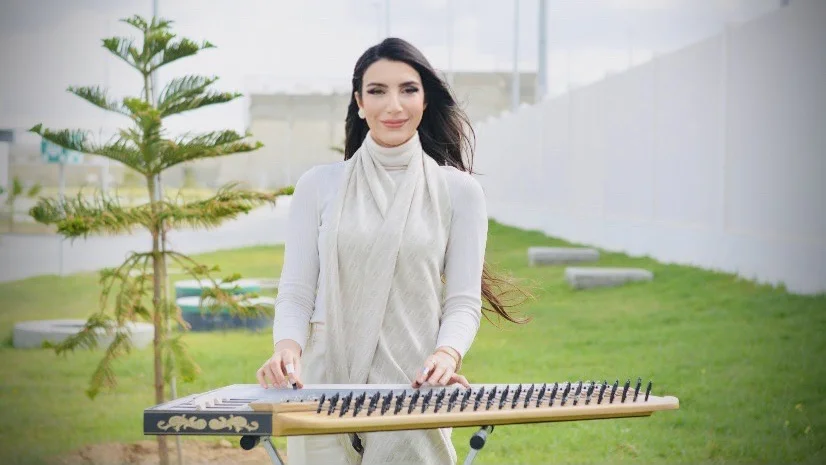 A woman in light-colored clothing plays the harp with a smile.